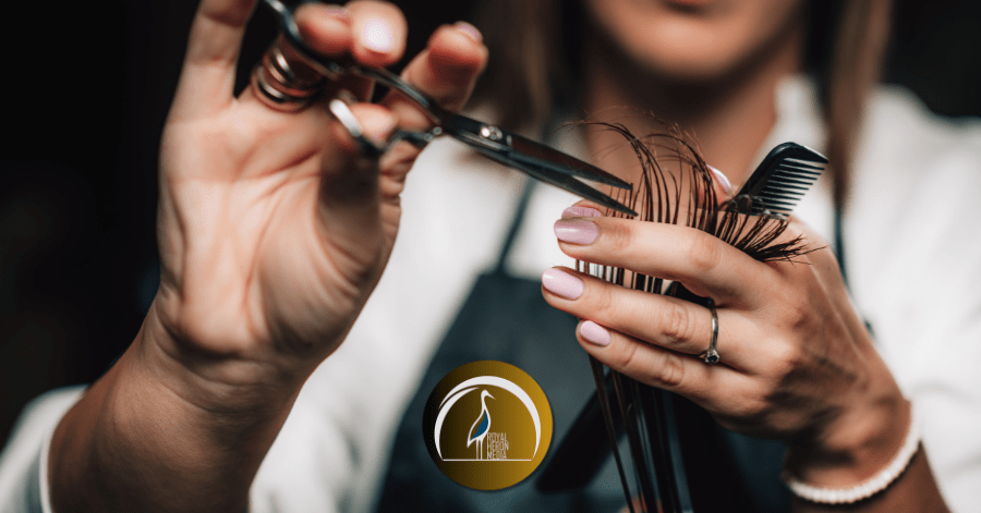 A professional hairdresser is cutting hair, holding scissors in one hand and a comb in the other, with a golden emblem visible on their apron.