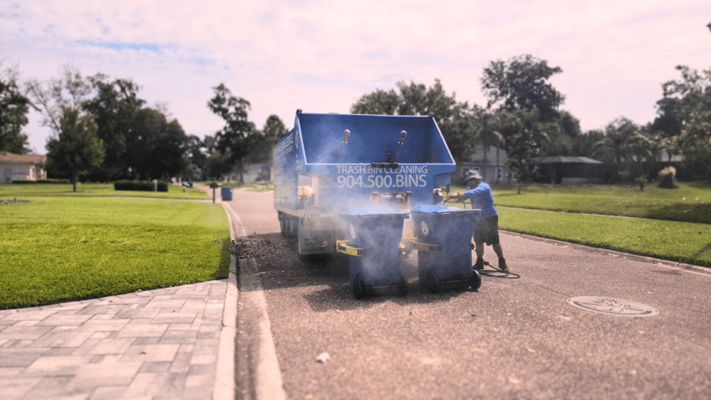 A person is cleaning trash bins using equipment connected to a blue truck labeled “TRASH BIN CLEANING” on a sunny day in a residential area