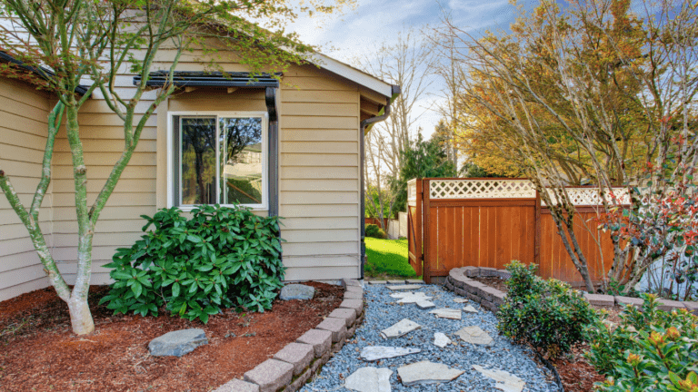 A peaceful backyard garden with lush plants, a stone pathway, and a wooden fence, located next to a beige house under a clear sky.